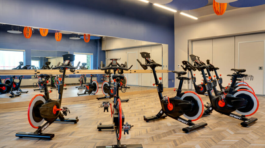 Fitness class room with chevron-designed floor, eliptical machines in foreground with back blue wall of mirrors and ballet bar. Ceiling has blue, beige, and orange hanging acoustic tiles.
