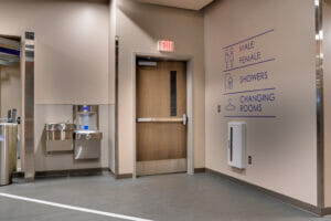 Interior gym view looking to a back exit door, right beige wall with blue vinyl directional decals and to the left a water fountain station.