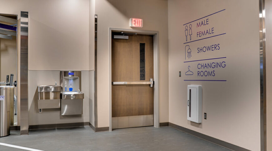 Interior gym view looking to a back exit door, right beige wall with blue vinyl directional decals and to the left a water fountain station.