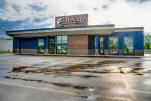 Exterior of wellness center with dark vibrant blue painted concrete on the upper part of the walls, with brown brick on the lowers and accents of brown wood paneling throughout. Then a "LifeCare Warroad Wellness" sign on the roof.