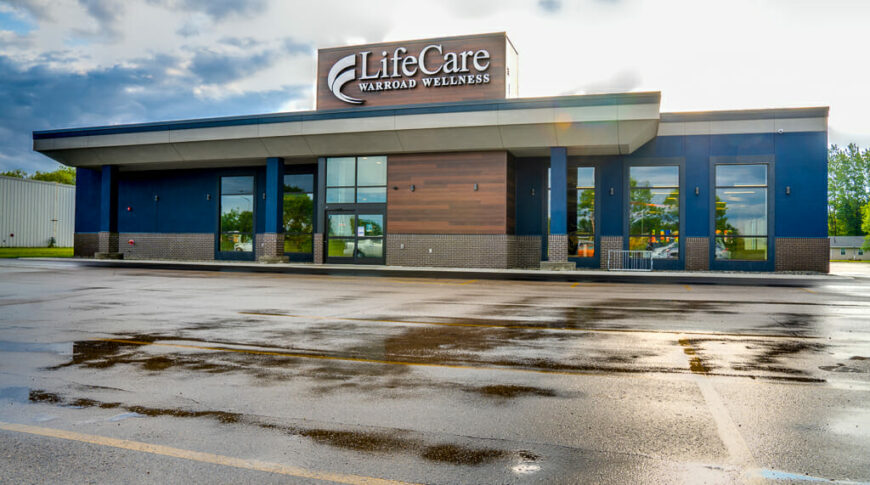 Exterior of wellness center with dark vibrant blue painted concrete on the upper part of the walls, with brown brick on the lowers and accents of brown wood paneling throughout. Then a "LifeCare Warroad Wellness" sign on the roof.