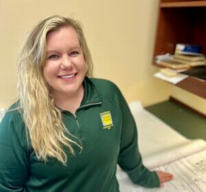 White woman with long bright blonde wavy hair in green long-sleeve Comstock Construction polo leaning next to desk with construction blueprints to pose for a picture that demonstrates the benefits of pre-construction services. phase.