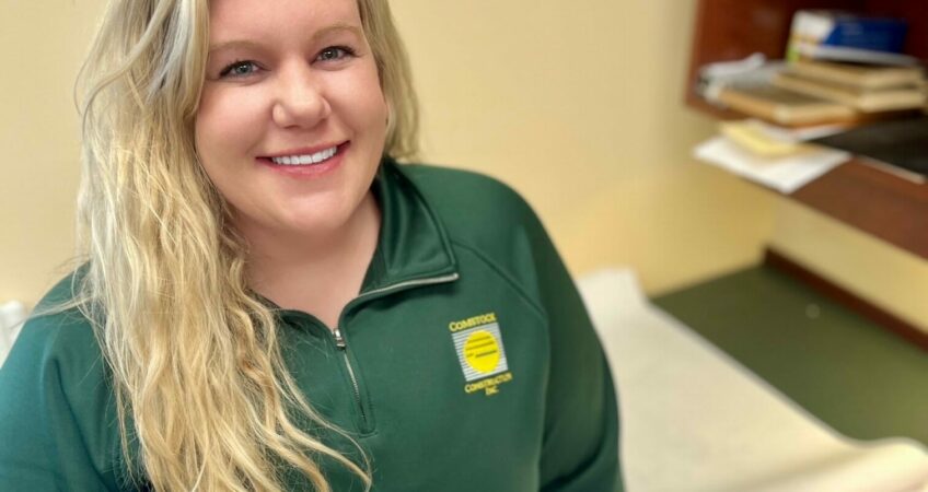 White woman with long bright blonde wavy hair in green long-sleeve Comstock Construction polo leaning next to desk with construction blueprints to pose for a picture that demonstrates the benefits of pre-construction services. phase.