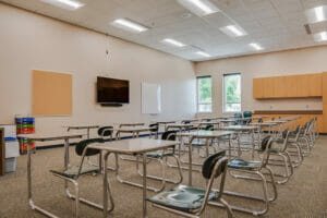 Beige-painted classroom with light maple cabinetry on the back wall alongside two tall windows, with desks in the middle of the room.