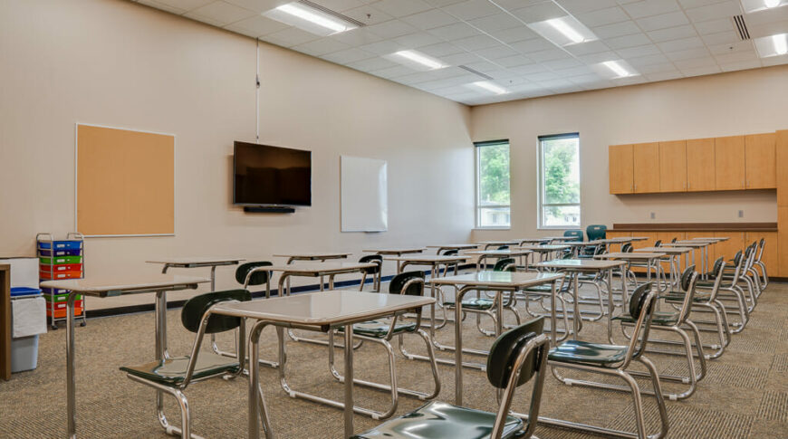 Beige-painted classroom with light maple cabinetry on the back wall alongside two tall windows, with desks in the middle of the room.