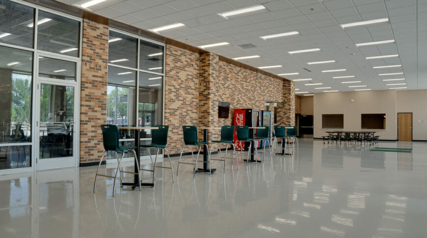 School commons area with light flooring with tall tables and chairs, vending machines in the background and brown brick wall behind.