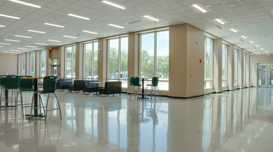 Corner view of school commons area with floor-to-ceiling windows overlooking parking lot and tables with booths and taller bar tables and stools throughout.
