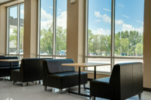 School commons area with tall double windows spaced every couple feet and tables and bench seats below.