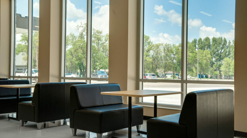 School commons area with tall double windows spaced every couple feet and tables and bench seats below.