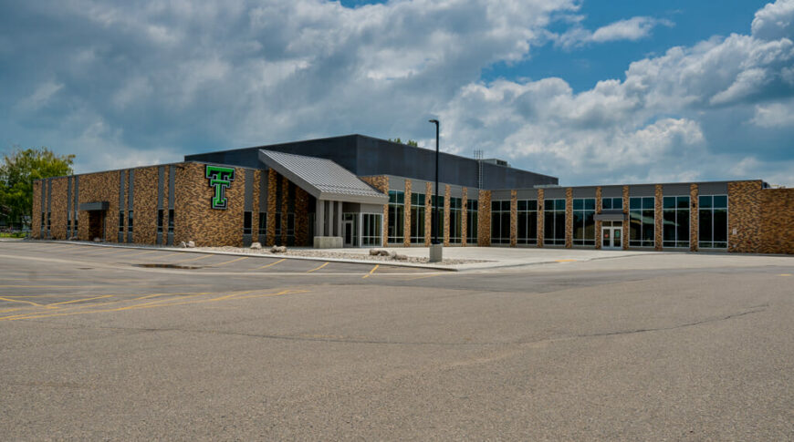 Exterior view of Thompson School addition with brown brick and gray metal exterior and green "TT" school marquee and parking lot to the foreground.