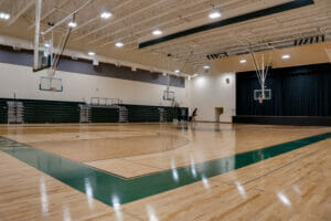 School basketball court with stacked bleachers to the back left wall and dark curtains covering stage to the back right wall.