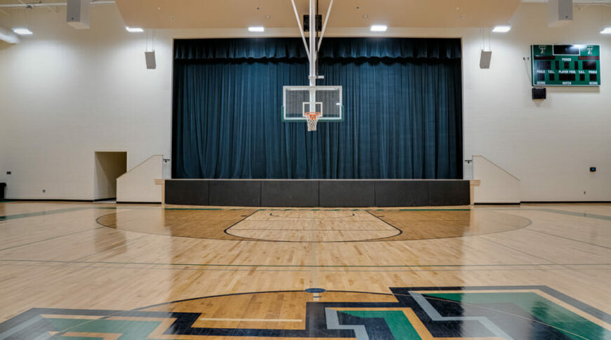 School gymnasium from the middle of the court looking ahead to a basketball hoop and dark draped stage behind.