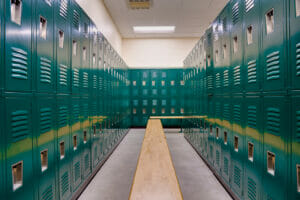 School gym locker room with double-stacked green lockers and light colored wooden bench seats underneath.