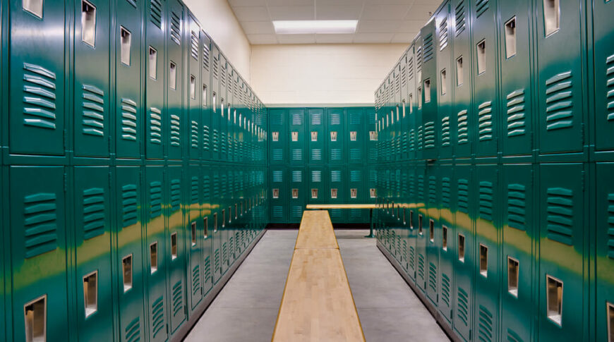 School gym locker room with double-stacked green lockers and light colored wooden bench seats underneath.