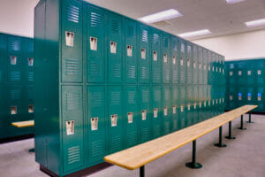 School gym locker room with double-stacked green lockers and light colored wooden bench seats underneath.