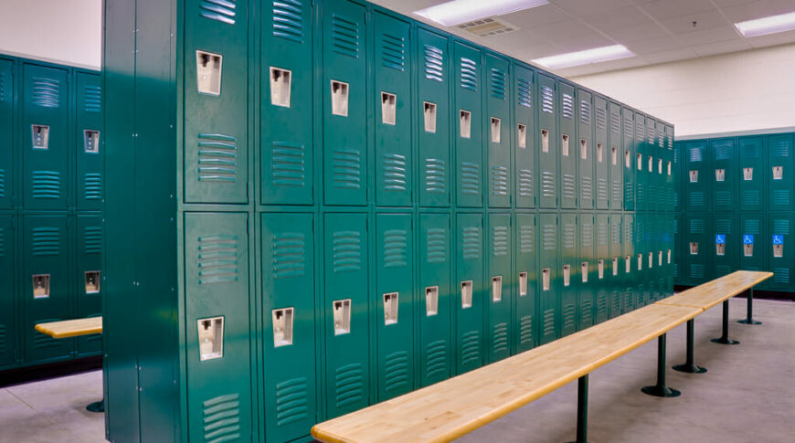 School gym locker room with double-stacked green lockers and light colored wooden bench seats underneath.