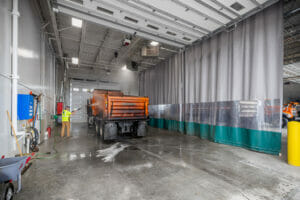 Wash bay side of the truck garages with man in yellow construction shirt washing an orange dump truck.