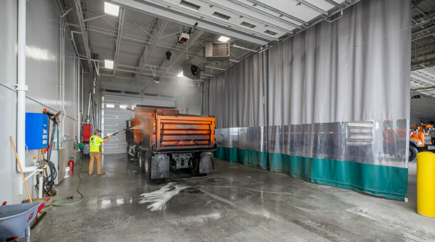 Wash bay side of the truck garages with man in yellow construction shirt washing an orange dump truck.