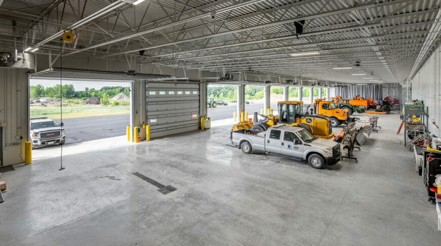 County truck garage bay from second floor looking down at concrete floor with trucks and heavy equipment with open garage bay doors in the background.