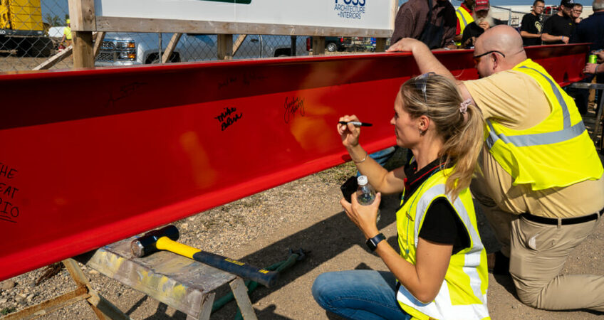 Woman and man in safety yellow vest kneeling down to sign their name on a red construction beam.
