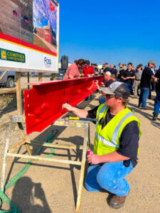 Employees kneeling down signing their names on a red structural beam waiting to be hoisted into place.