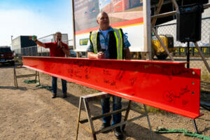 Two men standing behind red structural beam hoisted on two saw horses with construction sign in the background.