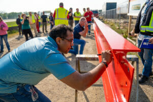 Man in blue polo signing red beam with sharpie. Group of people in the background watching.