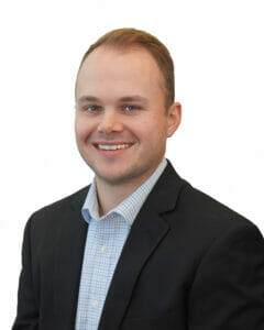 Young professional male with light skin, blue eyes, and dark blonde hair wearing black suit jacket and checkered blue grid collared shirt posing for professional portrait with white background.