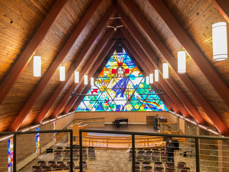 View from balcony of a-frame church overlooking chapel floor with wooden shiplap ceiling and triangular-shaped stained glass wall behind the pulpit.