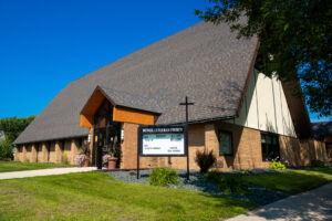 Exterior of steep A-framed Bethel Lutheran church building with dark brown shingles and light ornage bricks. Sign in front of gabled entrance.