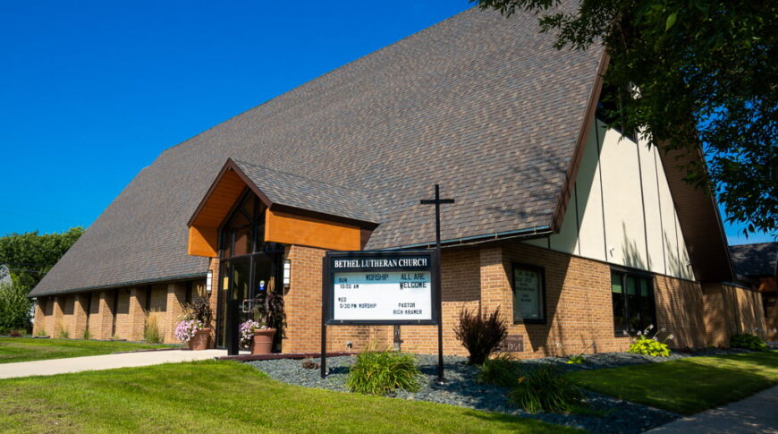 Exterior of steep A-framed Bethel Lutheran church building with dark brown shingles and light ornage bricks. Sign in front of gabled entrance.