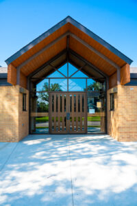 Gabled porch awning covering entrance to light orange bricked building. Double doors with glass surrounded windows.