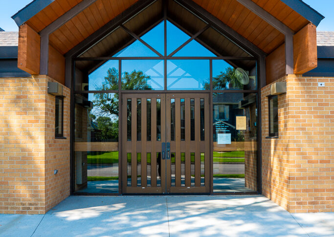 Gabled porch awning covering entrance to light orange bricked building. Double doors with glass surrounded windows.