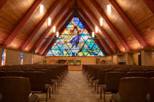 Interior of large A-framed Bethel Lutheran church sanctuary with large ceiling beams and wood paneling on the ceiling, with chairs underneath. Pulpit stage area to the back with a large stained glass religious window in the background.