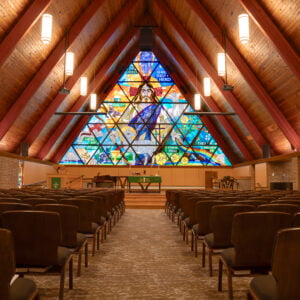 Interior of large A-framed Bethel Lutheran church sanctuary with large ceiling beams and wood paneling on the ceiling, with chairs underneath. Pulpit stage area to the back with a large stained glass religious window in the background.