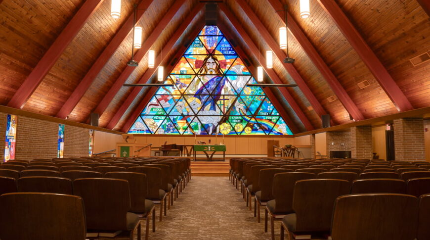Interior of large A-framed Bethel Lutheran church sanctuary with large ceiling beams and wood paneling on the ceiling, with chairs underneath. Pulpit stage area to the back with a large stained glass religious window in the background.