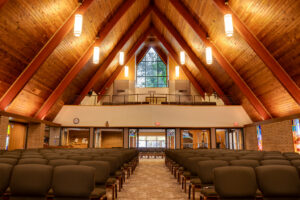 Interior of wooden and brick A-framed church sanctuary with a view from the pulpit looking out towards the crowd, with a choir balcony above.