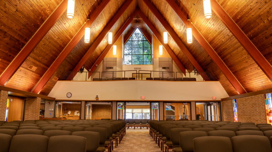 Interior of wooden and brick A-framed church sanctuary with a view from the pulpit looking out towards the crowd, with a choir balcony above.