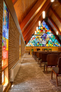 Side aisle view of Lutheran church sanctuary with vertical stained glass windows accenting the brick walls to the side, and to the back of the image is the pulpit and A-shaped stained glass window behind.