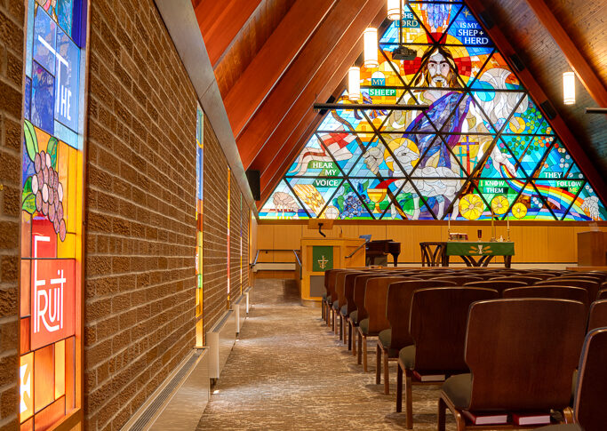 Side aisle view of Lutheran church sanctuary with vertical stained glass windows accenting the brick walls to the side, and to the back of the image is the pulpit and A-shaped stained glass window behind.