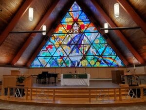 Interior view looking towards the honey oak chancel platform at a church with triangular-shaped stained glass window behind and a-frame wood paneled walls on either side.