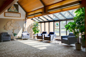 Interior large corridor of church building with wooden A-framed roof with a sunroom glass wall and ceiling to the right side and seating area throughout.