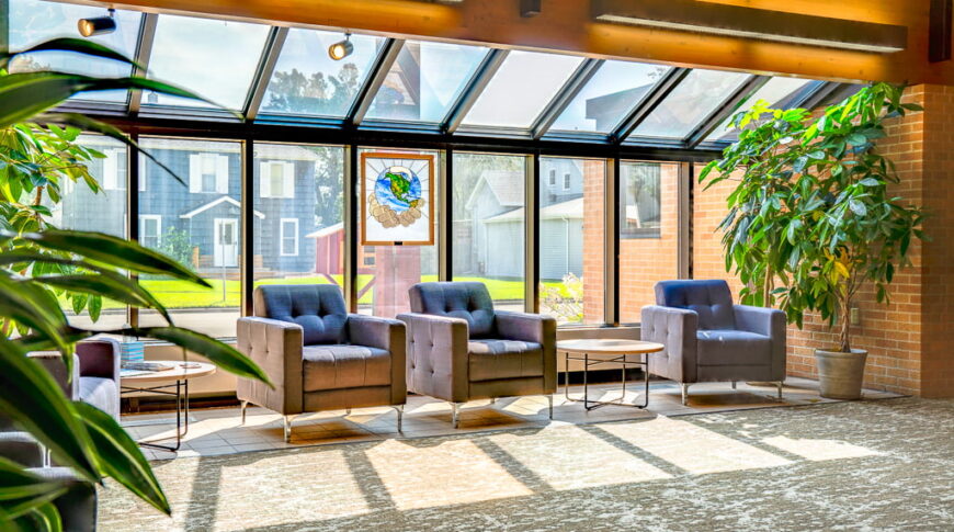 Atrium area of church hallway with jut-out glass walls and glass ceiling above. Seating area with chairs, tables, and plants underneath.