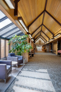 Long A-framed corridor of Bethel Lutheran church with wooden framed ceiling and beams above, gray carpet below and seating area on a bump-out to the left side.