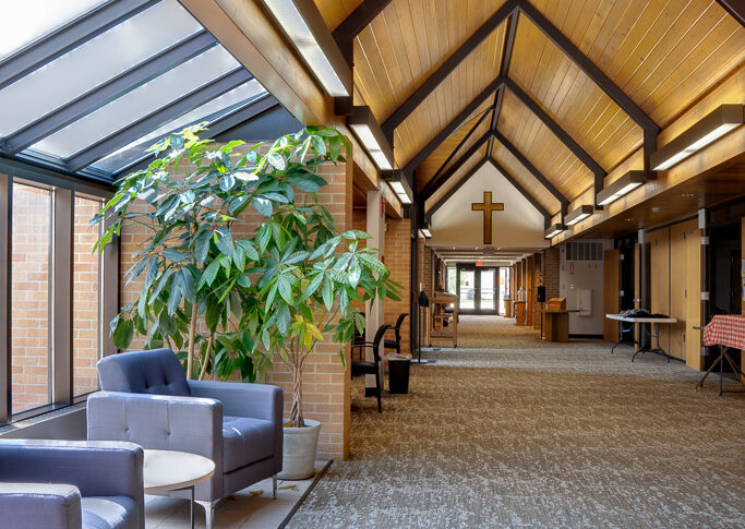 Long A-framed corridor of Bethel Lutheran church with wooden framed ceiling and beams above, gray carpet below and seating area on a bump-out to the left side.