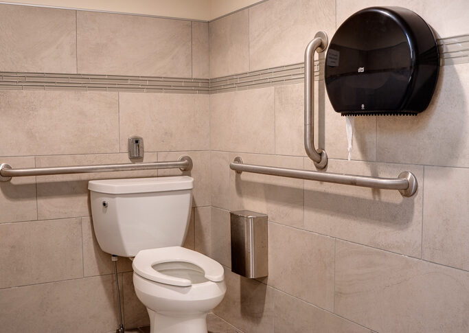 Interior of newly renovated ADA accessible bathroom with toilet, rai bars, receptacle and toilet paper dispensor.