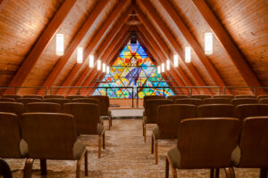 Choir balcony with cushioned chairs throughout overlooking a wooden A-framed ceiling and colorful stained-glass window behind the Bethel Lutheran Church pulpit.