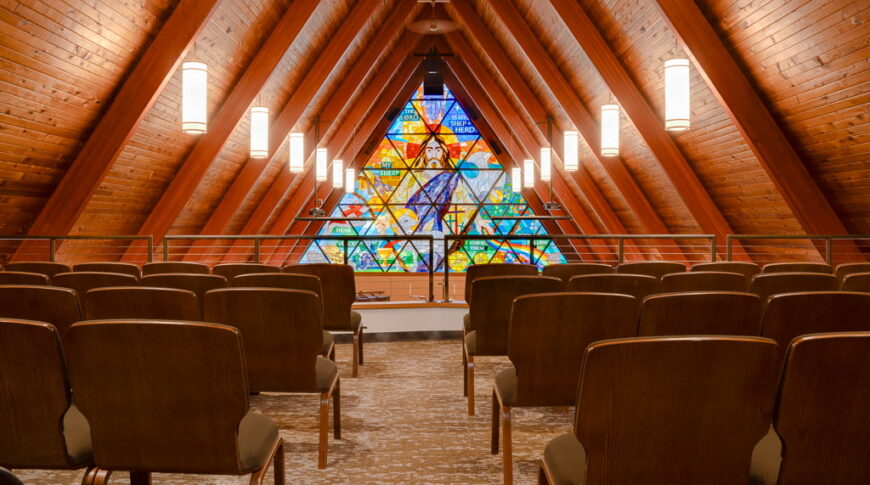 Choir balcony with cushioned chairs throughout overlooking a wooden A-framed ceiling and colorful stained-glass window behind the Bethel Lutheran Church pulpit.