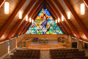 Choir balcony view looking down over the majestic wooden A-framed church sanctuary with cushioned chairs below, wooden pulpit at the front, with large framed stained glass window behind.
