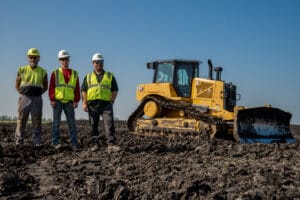 Three men with hard hats and safety vests stand for a picture in a dirt field with a bulldozer in the background.